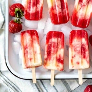 An image of two strawberry pineapple coconut swirl popsicles side by side on a bed of ice cubes.