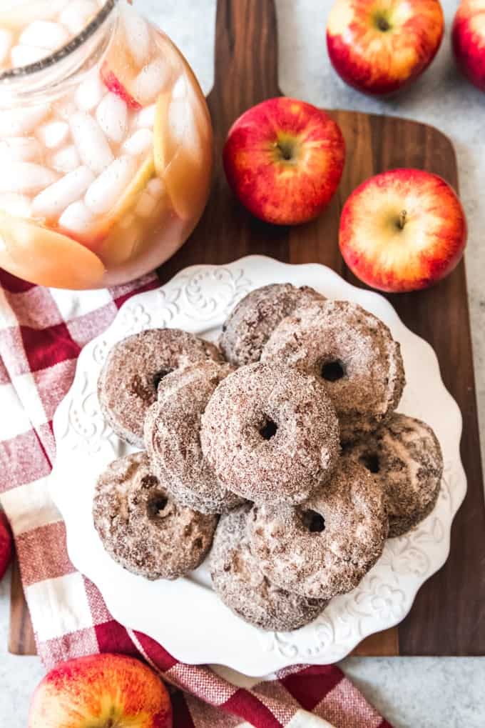 An image of a stack of apple cider donuts with a pitcher of apple cider and apples surrounding it.