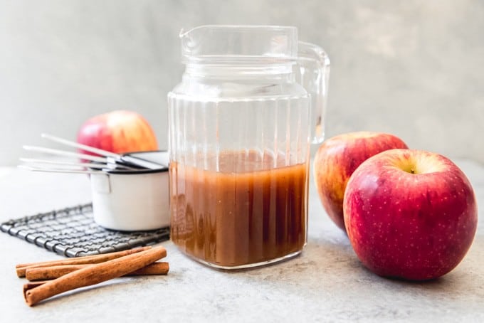 An image of a glass jar of homemade cider syrup with cinnamon sticks and apples next to it.