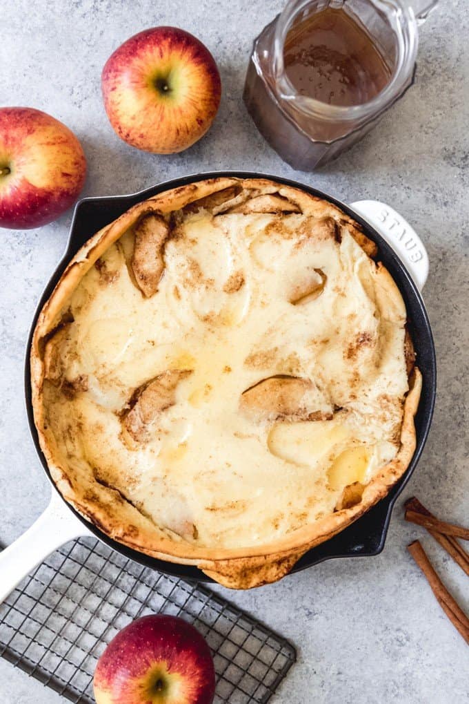 a fluffy apple german pancake in a skillet with fresh whole apples and syrup to the sides