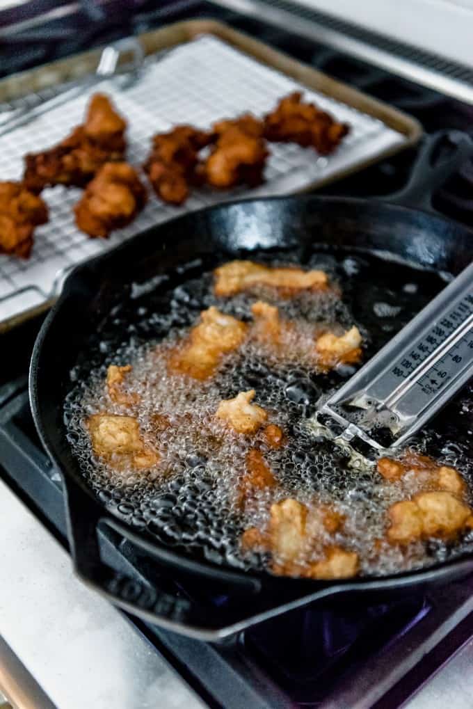 a cast iron skillet with fish frying and a thermometer inside and fried fish in the background on a wire rack