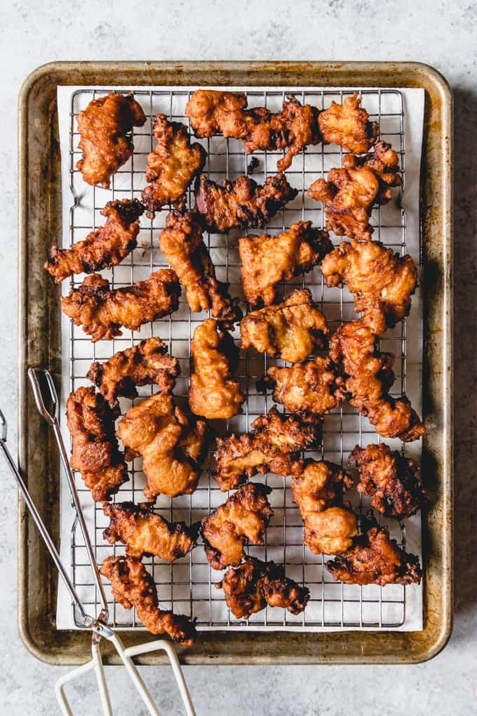 fried fish on a wire rack on a paper towel lined baking sheet with metal tongs