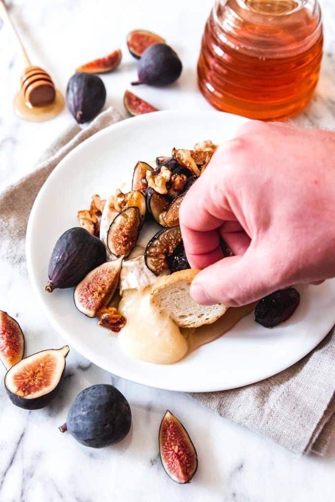 An image of a hand holding a slice of toasted baguette scooping up melted baked brie cheese with figs, walnuts, and honey.