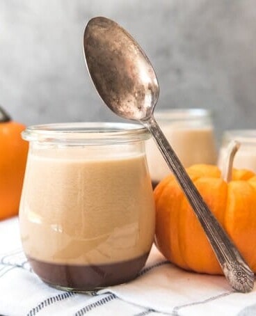 a glass jar filled with pumpkin panna cotta with fresh small pumpkins and a spoon around it