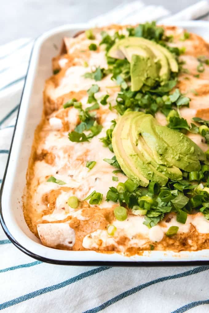 An image of homemade chicken enchiladas with pumpkin enchilada sauce, topped with chopped green onions, cilantro, and avocado.