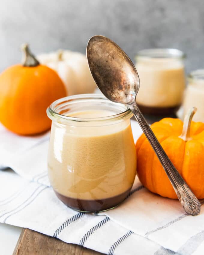 A spoon resting off of the glass jar of caramel panna cotta next to a small pumpkin.