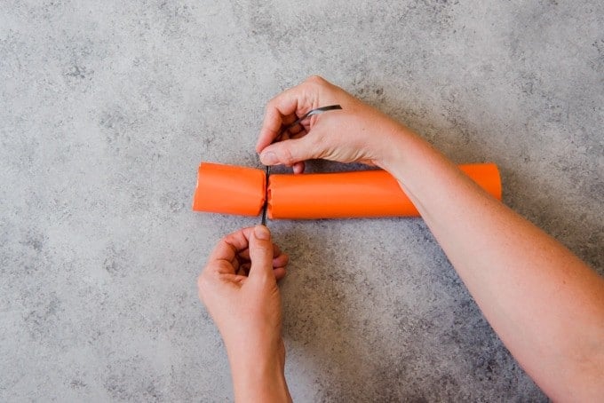 An image of hands tying black curling ribbon around an orange colored Christmas cracker for Halloween.