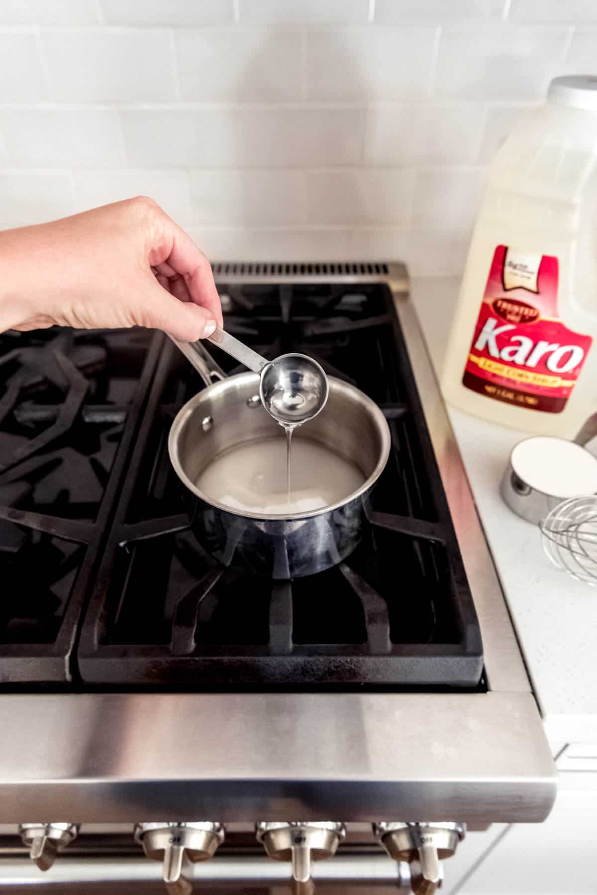 An image of a saucepan on a stove with a hand holding a tablespoon of corn syrup pouring in ingredients for salted caramel sauce.