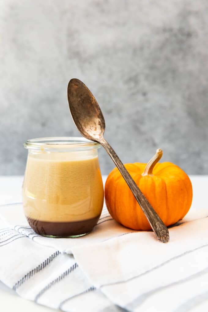 A spoon resting off of the glass jar of caramel panna cotta next to a small pumpkin.