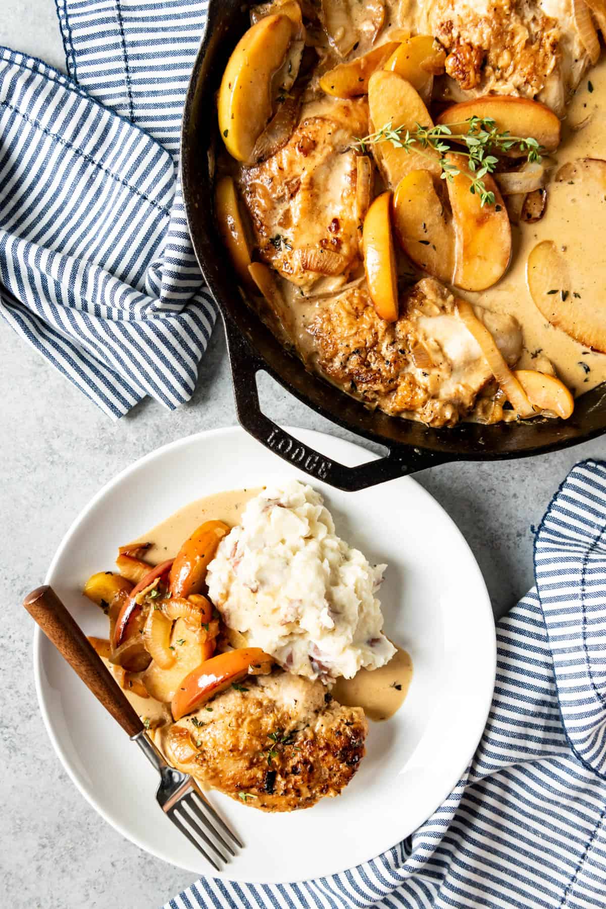 A plate of mashed potatoes with creamy apple cider chicken next to a large skillet of chicken and apples.