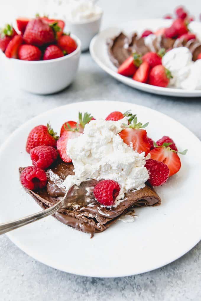 An image of chocolate crepes filled with nutella and topped with freshly whipped cream, raspberries and strawberries.