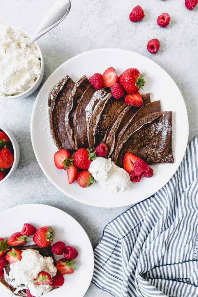 An image of stacked chocolate crepes on a plate with fresh strawberries and raspberries for filling the crepes.