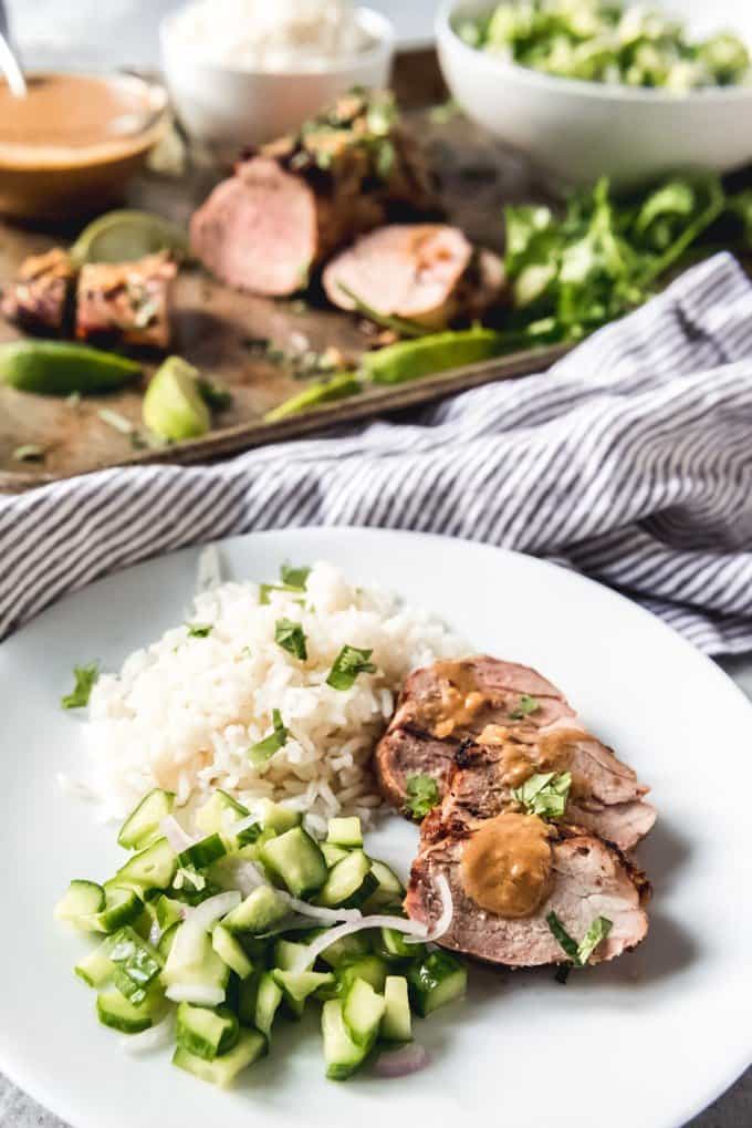 An image of a plate of sliced pork tenderloin topped with a Thai peanut sauce, served with spicy cucumber salad and coconut rice on the side.