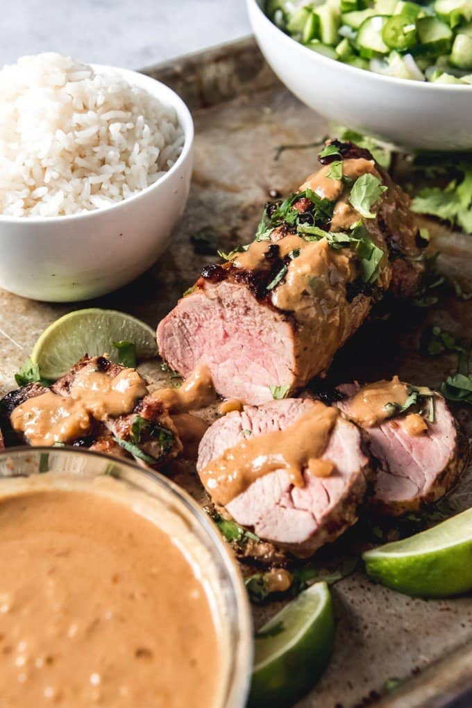An image of a grilled pork tenderloin sprinkled with cilantro and an easy peanut sauce, surrounded by extra sauce, a bowl of rice, and a cucumber salad.