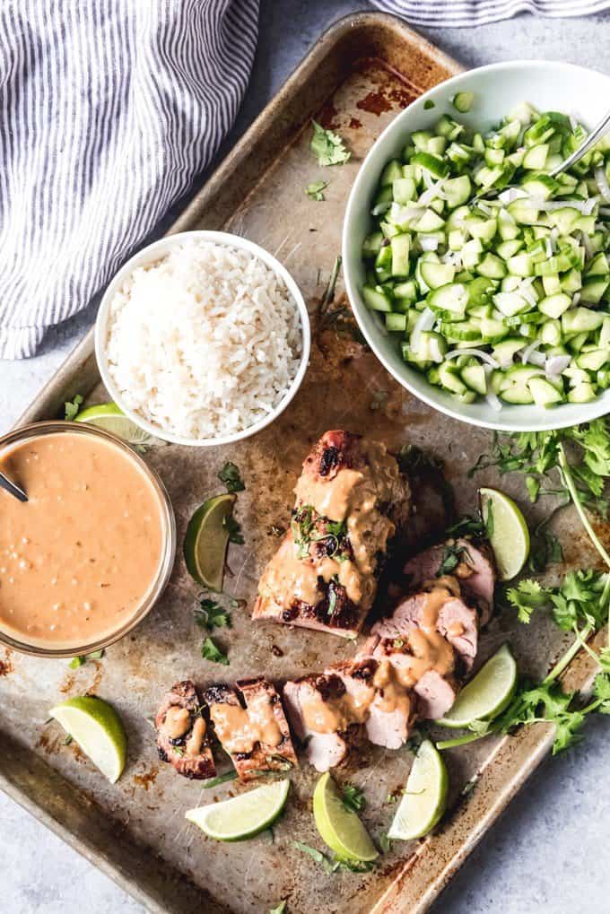 An image of a grilled Thai pork tenderloin with spicy peanut sauce, coconut rice, and cucumber salad on the side.