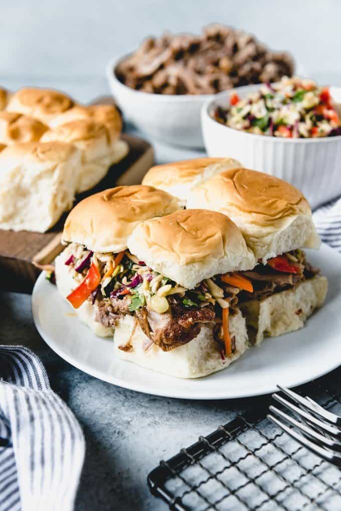 asian pulled pork sliders with asian slaw on a white plate in front of more buns, pulled pork, and asian slaw