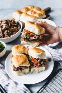 a hand holding a slow cooked asian pulled pork sandwich over a plate of sandwiches