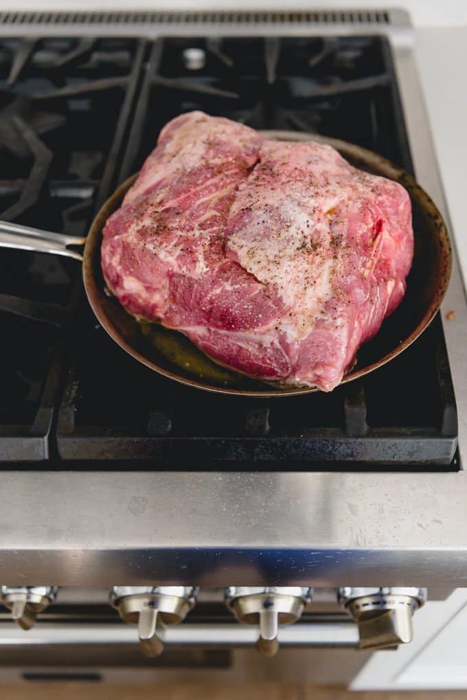 An image of a bone-in pork shoulder butt roast seasoned with salt and pepper, searing on a hot stove in a pan.