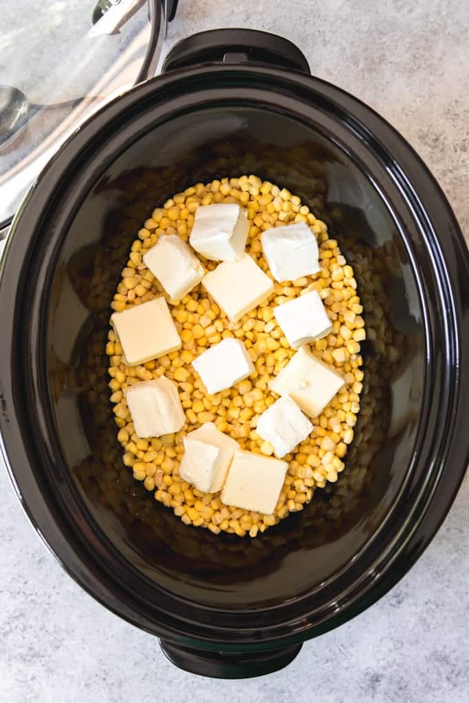 An image of a crock pot with frozen corn, cubed butter, and cream cheese for slow cooker creamed corn.
