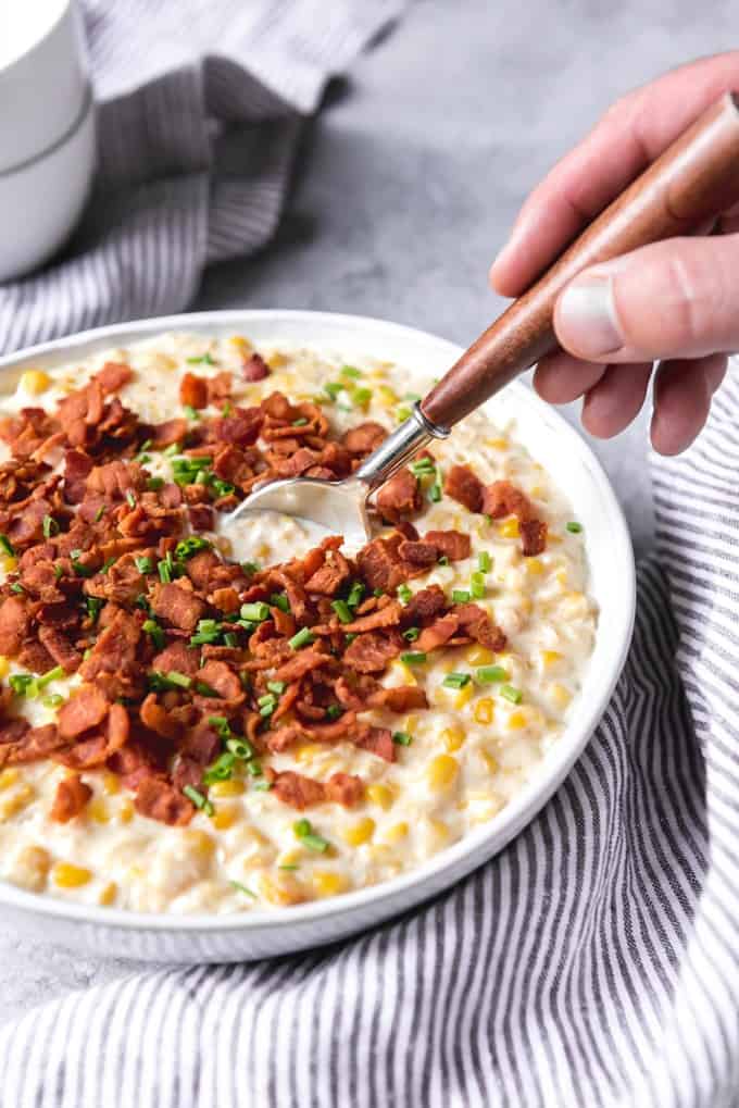 Creamed corn topped with bacon in a bowl with a hand sticking a spoon into it.