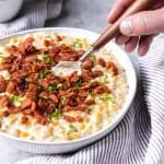 a hand dipping a spoon into a bowl of creamed corn with bacon bits on top