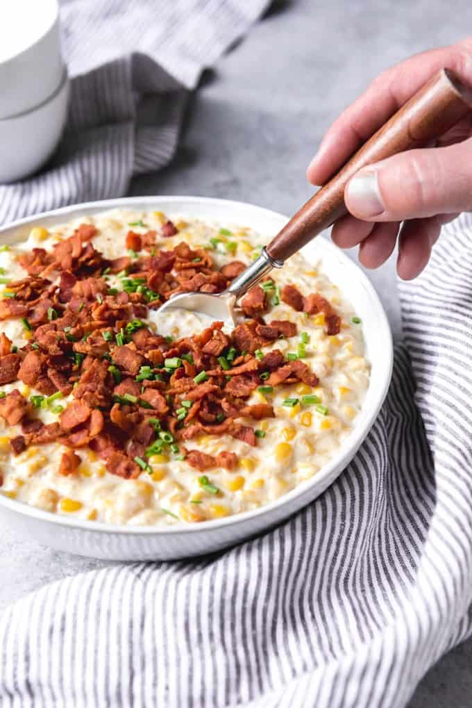 A white bowl filled with creamed corn, topped with crispy bacon bits and chopped chives, with a spoon in it. 