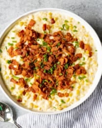 A large serving bowl of creamed corn with bacon next to a striped cloth napkin and spoon.