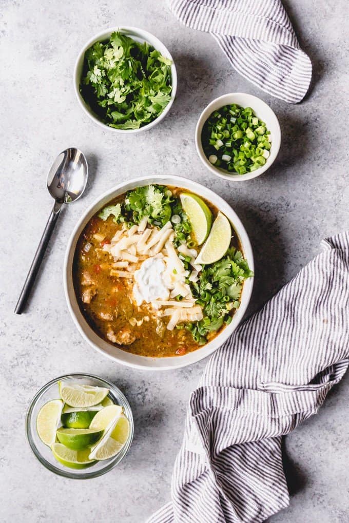 An image of a bowl of slow cooker chili verde made with chunk of tender pork, then topped with sour cream, cheese, cilantro, and lime wedges.