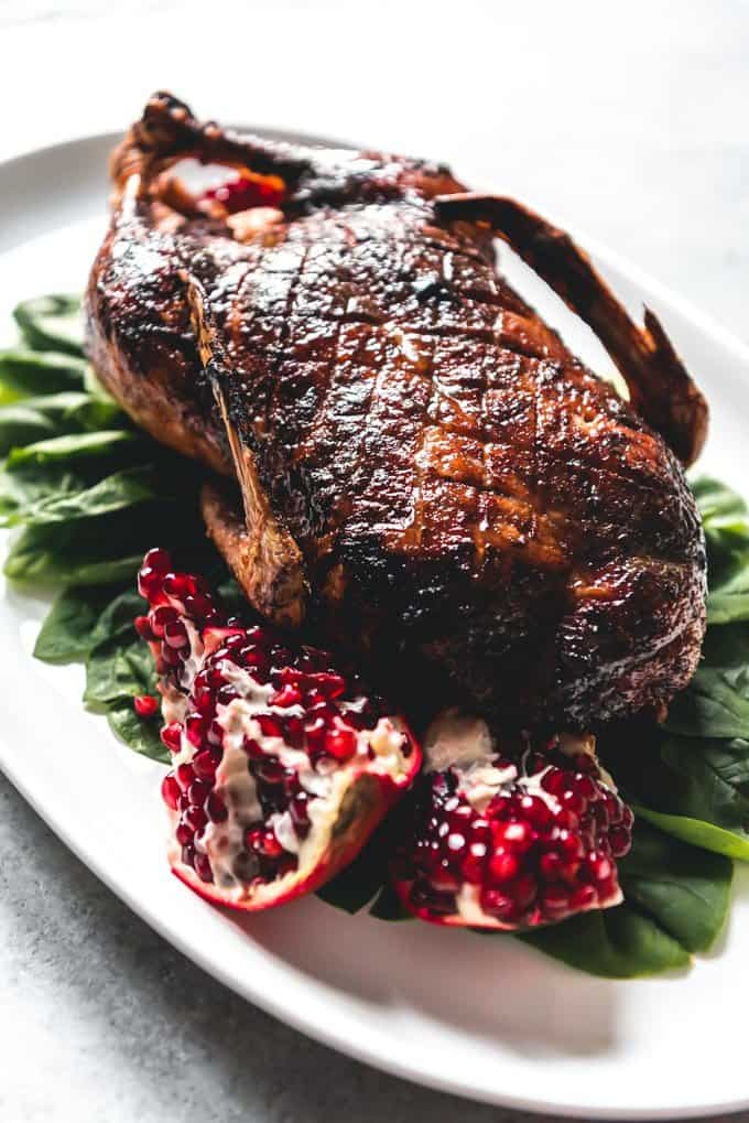 An image of a whole honey roast duck on a bed of spinach leaves on a white serving platter, with pomegranate seeds around it for presentation.