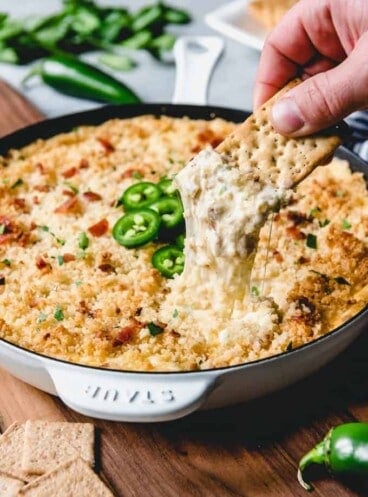 a cracker pulling some cheesy jalapeno dip out of a skillet