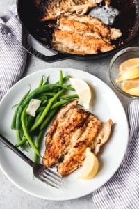 pan fried trout fillet on a plate with lemon wedges and green beans with more in a cast iron skillet to the side