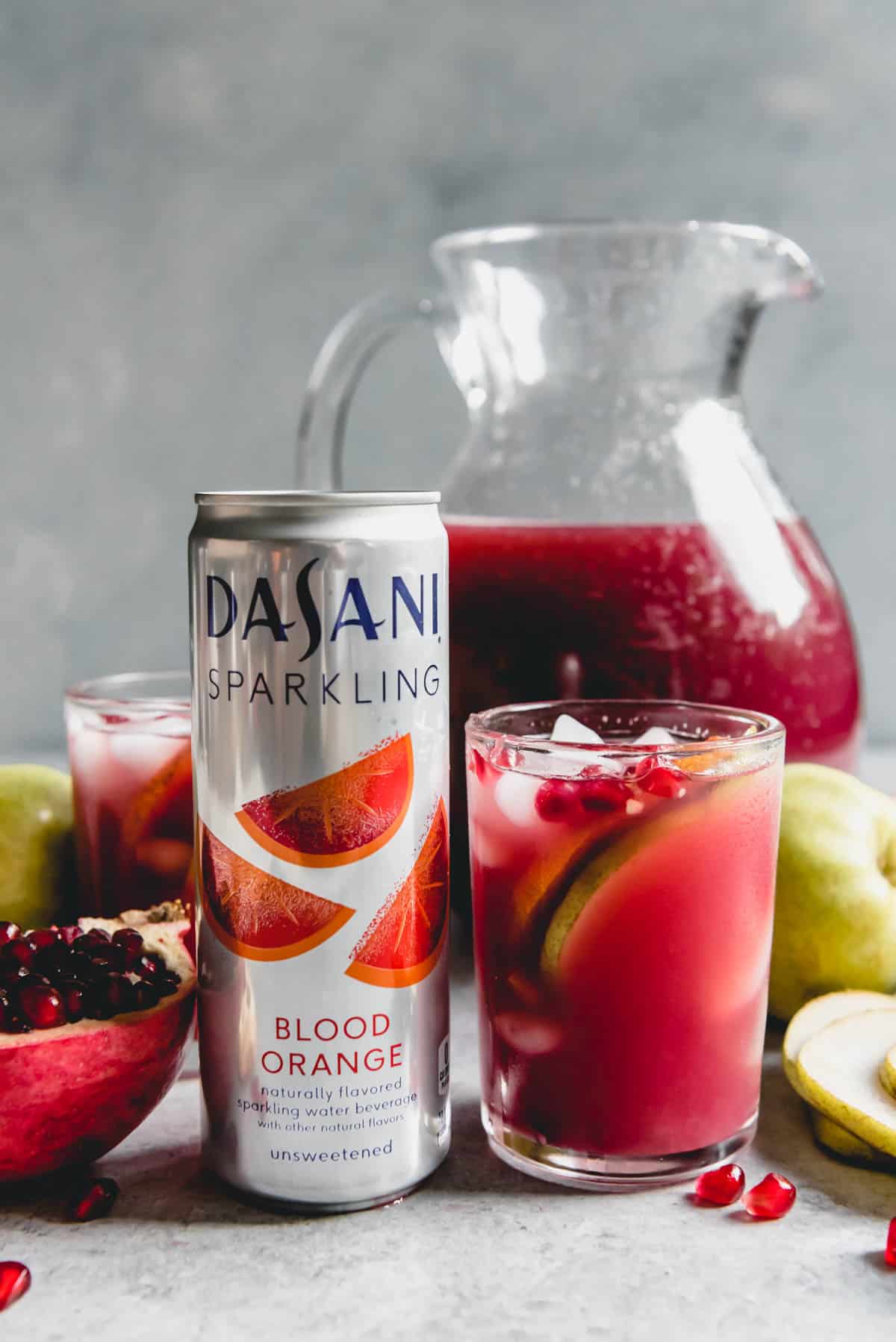 An image of a can of DASANI Sparkling Blood Orange flavored water beside a glass of Pomegranate in a Pear Tree Punch made with the flavors of blood oranges, pomegranate juice, and pear juice, then garnished with fruit.