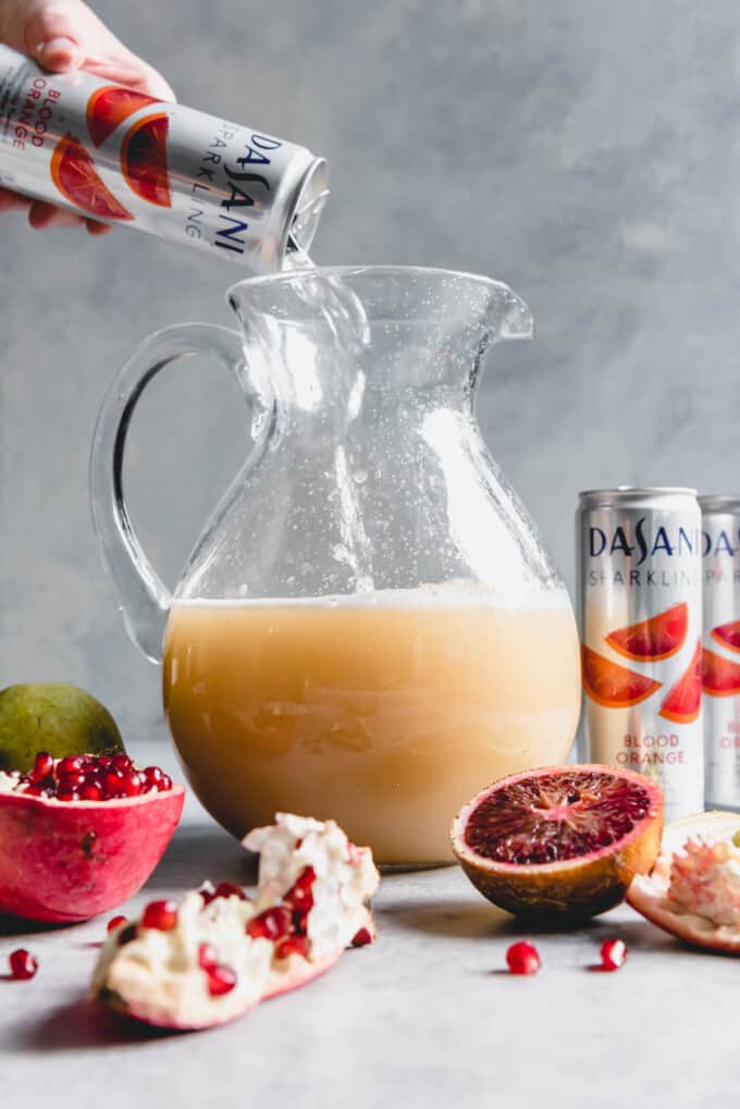An image of a glass pitcher partly filled with pear juice and a hand pouring a can of DASANI Sparkling Blood Orange flavored water into the pitcher.