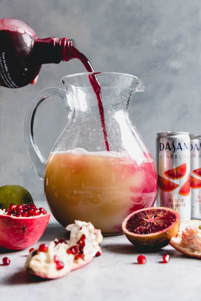 An image of pomegranate juice being poured into a tall glass pitcher along with pear juice and blood orange sparkling water to create a pomegranate in a pear tree punch for a festive holiday party.