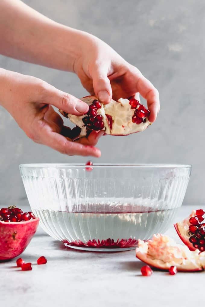 An image of hands opening a pomegranate and removing the seeds over a bowl of water showing how to cut open a pomegranate easily and without mess.