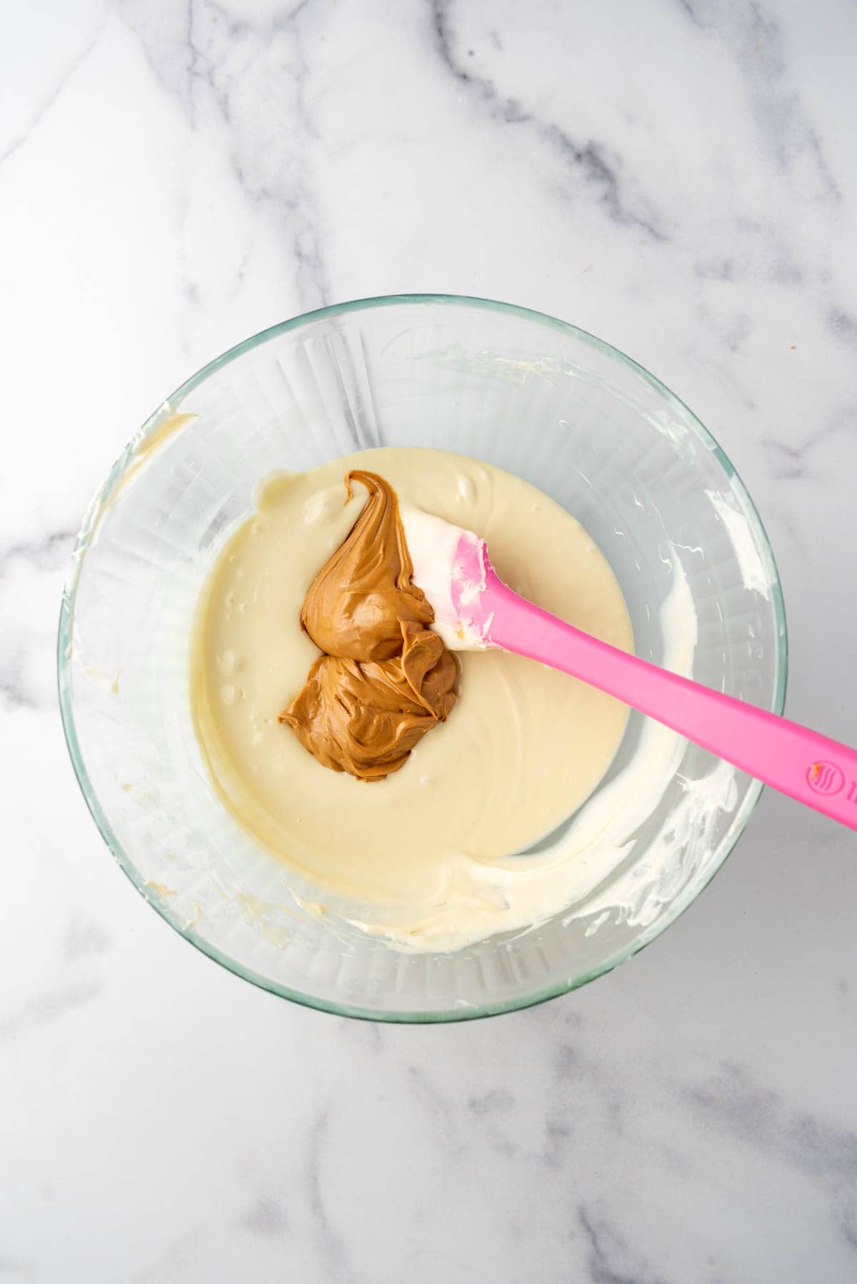 An image of creamy peanut butter being added to melted white chocolate in a large glass mixing bowl with a pink spatula.