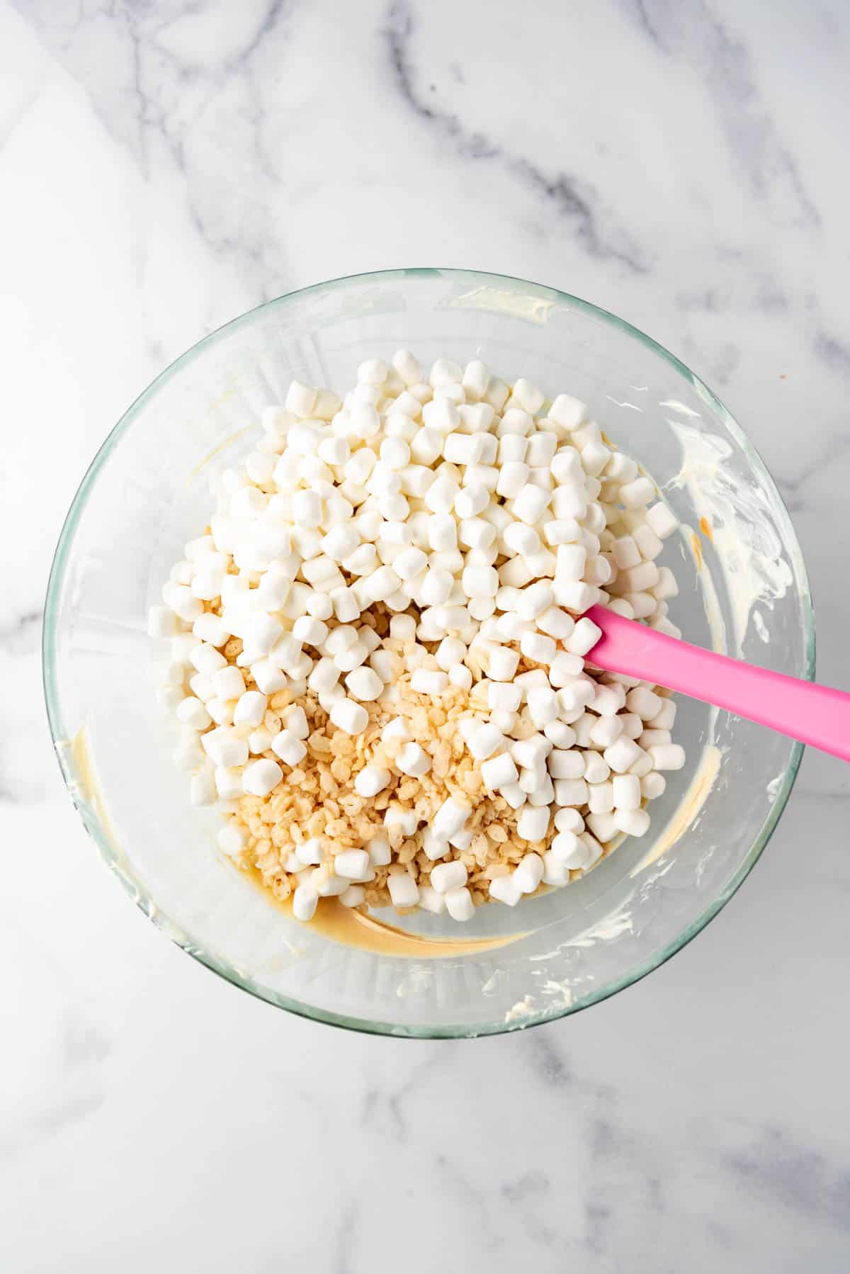 An image of mini marshmallows and rice krispies cereal being added to a mixing bowl of melted white chocolate and peanut butter.