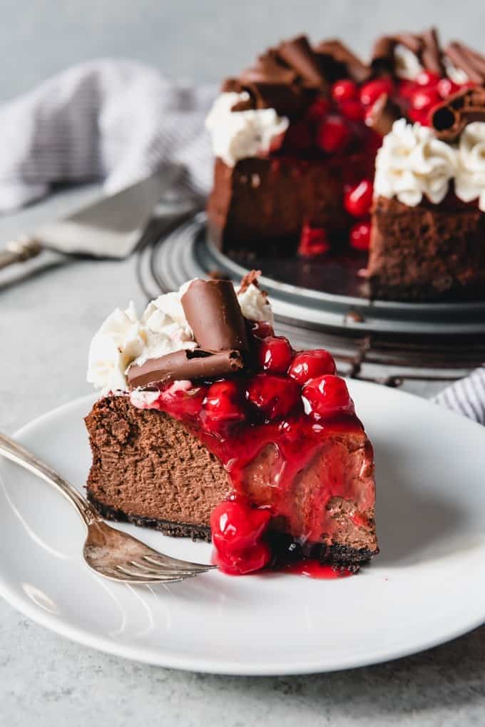 black forest cheesecake with cherry topping on a white plate with a fork with the rest of the cheesecake in back