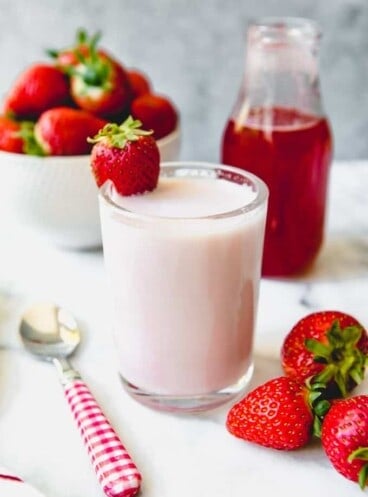 a cup of homemade strawberry milk with a glass of strawberry syrup in back and fresh strawberries to the sides