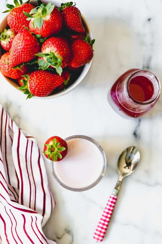An image of a bowl of strawberries next to a jar of strawberry syrup and a glass of strawberry milk.