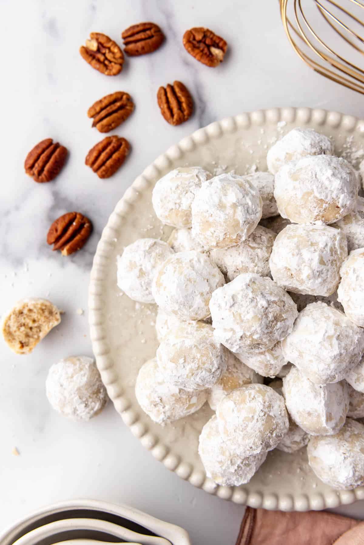 Powdered sugar covered cookies on a white plate next to scattered whole pecans.