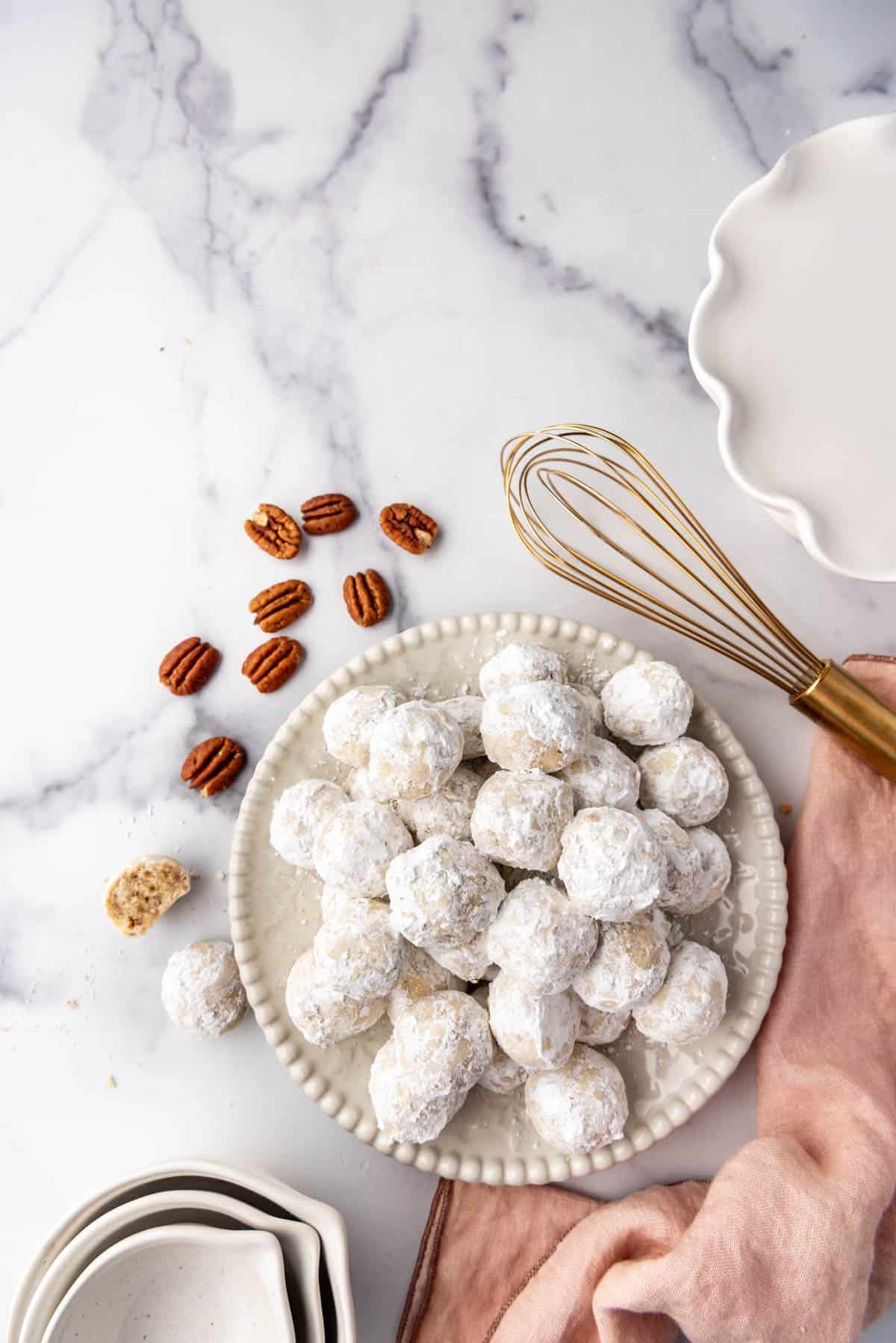 An overhead image of a plate of Mexican wedding cookies next to a pink cloth, whisk, and pecans.