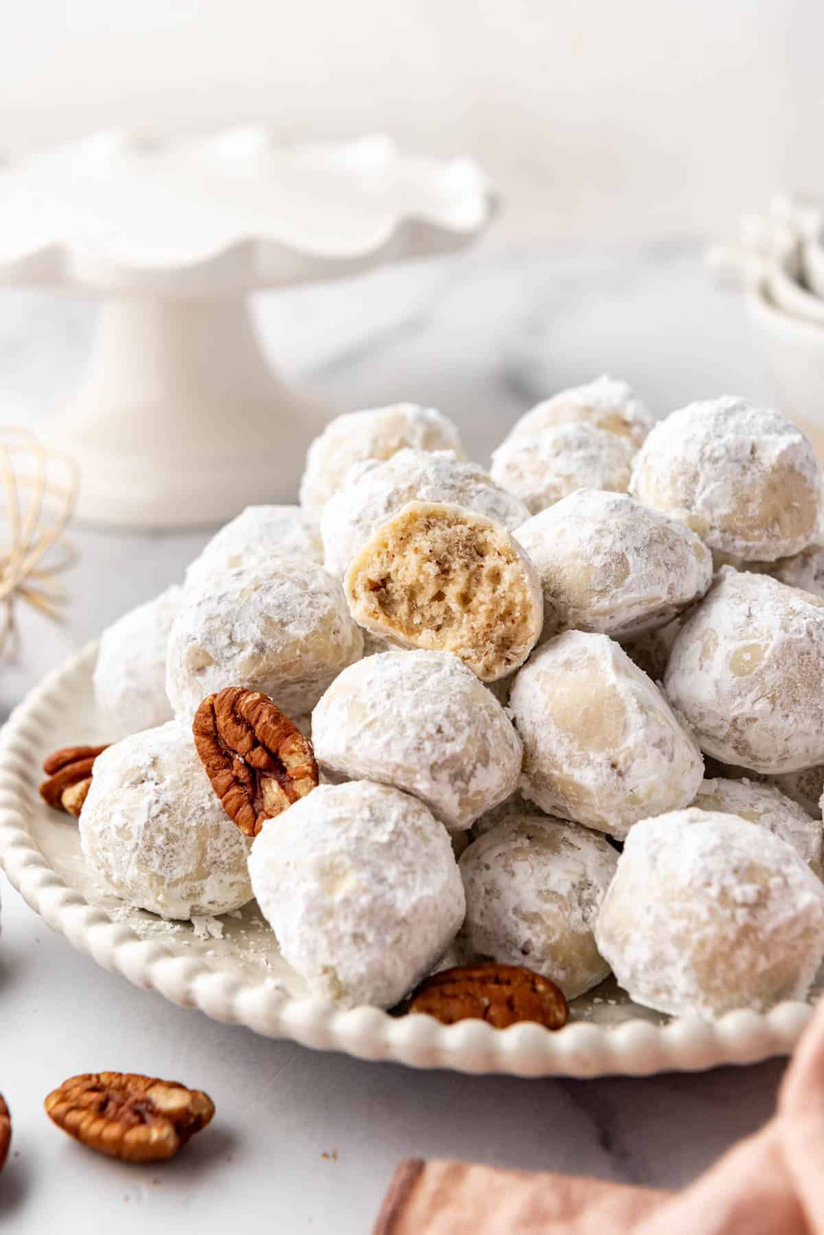 A pile of Mexican wedding cookies and pecans with one cookie bitten to reveal insides.