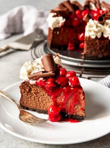 black forest cheesecake with cherry topping on a white plate with a fork with the rest of the cheesecake in back