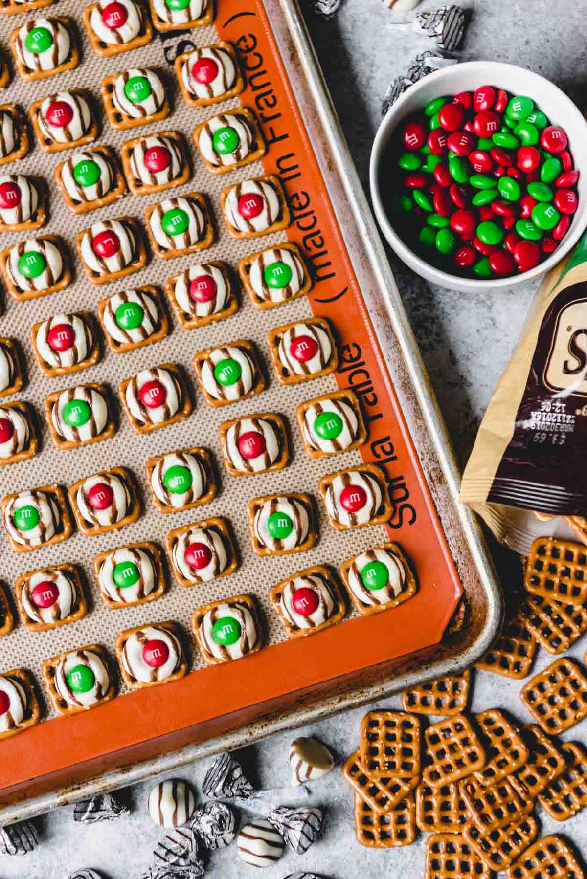 An image of bite-size Christmas treats made festive with red and green M&M's pressed into melted Hershey's hugs on top of square pretzels.