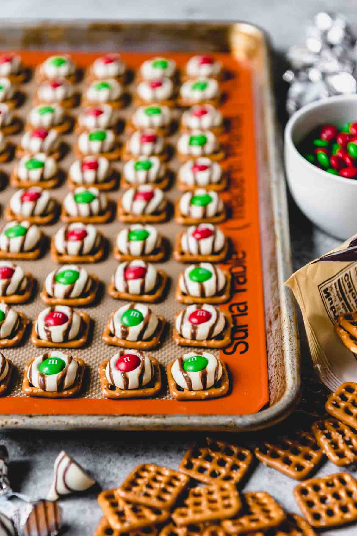 An image of easy pretzel hugs with red and green Christmas M&M's on a baking sheet with a silpat mat.