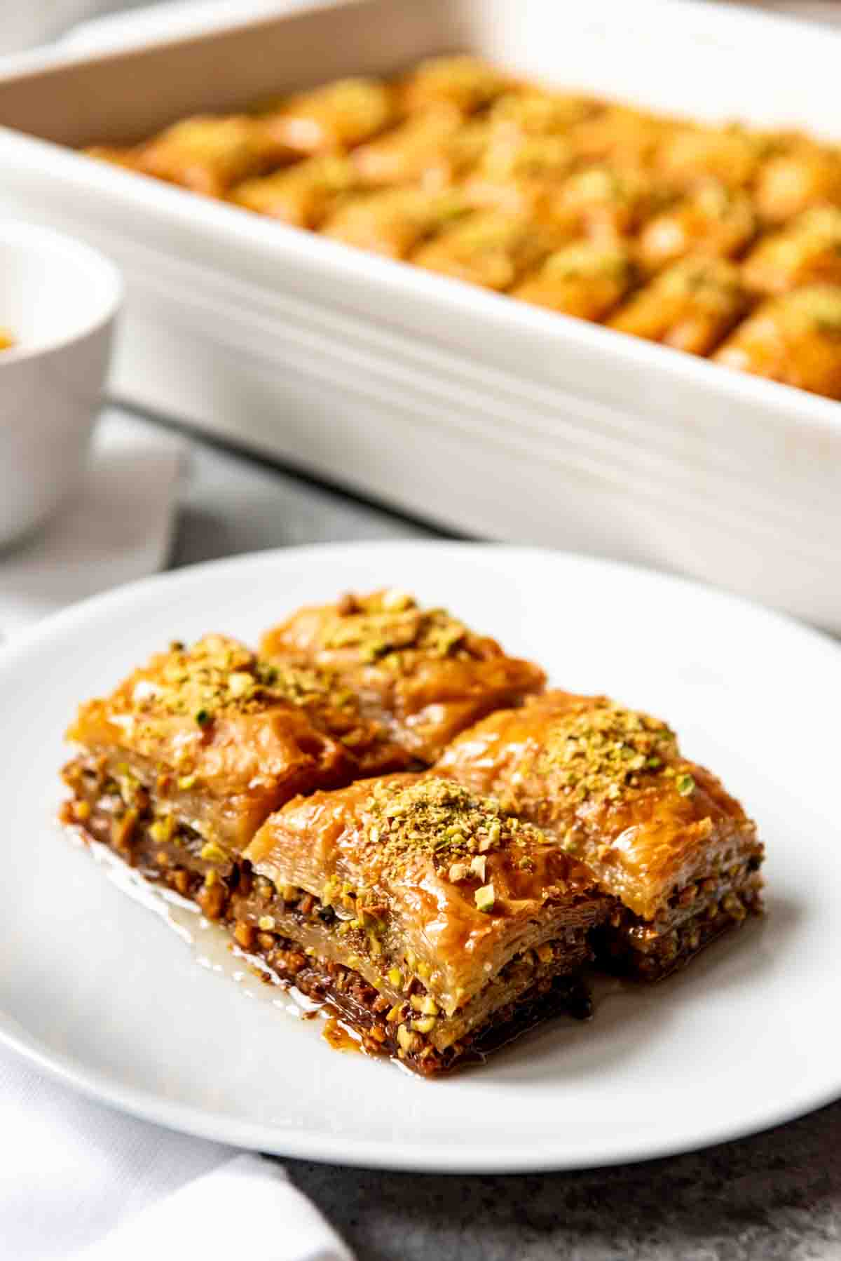 An image of fistikli baklava (Turkish baklava) made with pistachios, on a white plate in front of a pan full of baklava.