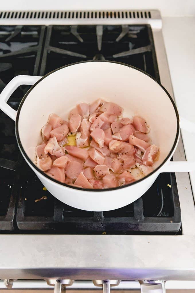 An image of bite-size pieces of chicken in a white dutch oven on a stove, being browned in just a little olive oil.