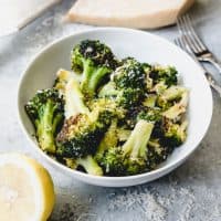 a white bowl with broccoli and cheese and half a lemon in front with a brick of parmesan cheese in back