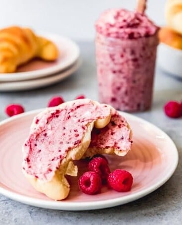 a white plate with a screscent roll split open and spread with raspberry butter on it and fresh raspberries to the side and scattered berries around the plate with a jar of butter and a plate and bowl of cresent rolls in the background